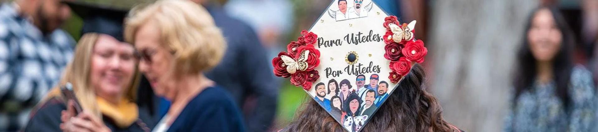 Student in regalia with a cap that reads "Para Ustedes, Por Ustedes" (For You, Because of You) with illustrations of friends and family
